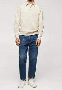 Sudadera beige clara con cuello, combinada con jeans azul medio y zapatillas blancas; ajuste relajado y diseño casual.