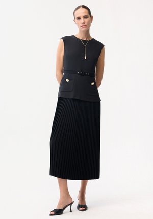 Kobieta stoi na białym tle, ubrana w bezrękawnikowy czarny top z paskiem i złotymi guzikami, plisowaną czarną midi spódnicę, czarne mule na obcasie oraz złoty naszyjnik.