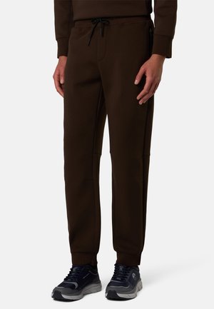 Persona che indossa pantaloni jogger marrone scuro con polsini elastici e scarpe da ginnastica nere su uno sfondo chiaro uniforme.