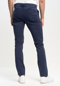 Temno modri usnjeni chinos hlače slim-fit kroja z gladko teksturo, ki imajo dva zadnja žepa in pas z zankami za pas. Kombinirano z belimi športnimi copati.