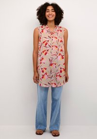 Mouwloze tuniek in zacht beige met roze en rode bladdessins, gedetailleerde halslijn; gecombineerd met lichtblauwe flared jeans en bruine sandalen.