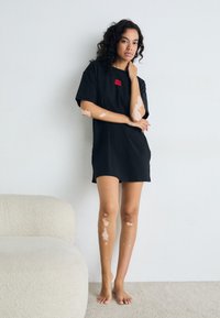 Juoda per didelis t-shirt su raudonu logotipo pleistru, trumpomis rankovėmis, apvaliu iškirpimu ir šoninėmis skląstėmis. Nešiojama basa prieš neutralų foną.