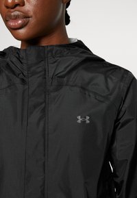 Gros plan d’une personne portant une veste à capuche noire Under Armour avec une fermeture éclair à l’avant et un logo sur la poitrine.