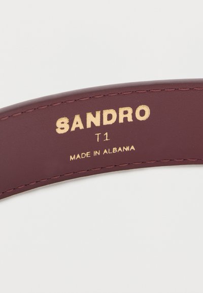 Κοντινή λήψη μαρον δερμάτινου αντικειμένου με χρυσό κείμενο "SANDRO T1 MADE IN ALBANIA" κεντημένο κατά μήκος της άκρης.