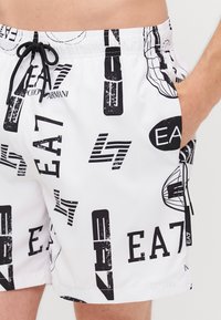 Pantaloni da bagno bianchi con stampe grafiche nere e testo, caratterizzati da una vita elastica e cordino. Due tasche laterali.