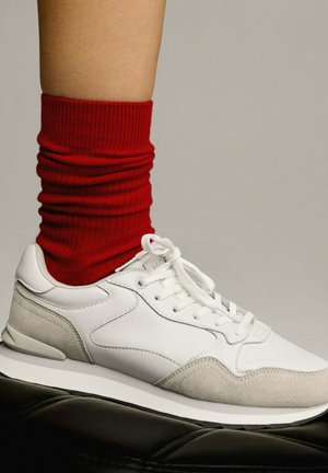Witte sportieve sneakers met grijze suède accenten, veters en een gevoerde zool, gecombineerd met een rode geribbelde sok.