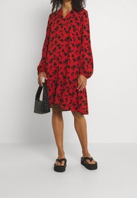 Vestido floral rojo con mangas largas y escote en V, con un dobladillo en capas. Combinado con un bolso negro trenzado y sandalias negras de suela gruesa.