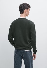 Donkergroene gebreide trui met een ronde hals en lange mouwen, gecombineerd met grijze spijkerbroek. De textuur oogt zacht en casual.