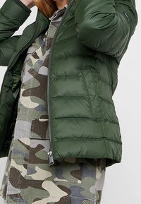 Grön puffjacka med en glänsande finish, med kviltade paneler och dragkedjestängning, som bärs tillsammans med en skjorta med camouflage-mönster under.