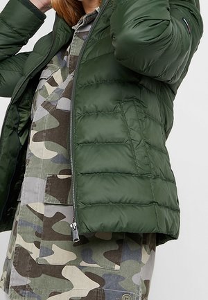 Grüne Pufferjacke mit glänzender Oberfläche, mit gesteppten Einsätzen und Reißverschluss, kombiniert mit einem Camouflage-Shirt darunter.