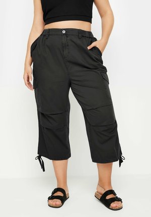 Woman wearing black cropped cargo pants with side pockets and black sandals, standing against a white background.