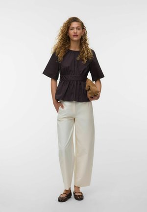 Vrouw met krullend haar die een zwarte blouse met korte mouwen draagt, witte broek met wijde pijpen, zwarte gevlochten platte schoenen, en een bruine clutch vasthoudt.