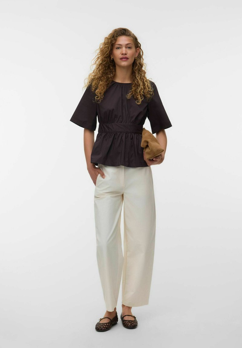 Femme aux cheveux bouclés portant un chemisier noir à manches courtes, un pantalon blanc à jambes larges, des ballerines noires tissées, tenant une pochette marron.