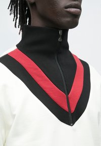 Valge kõrge kaelusega dressipluus, millel on must soonikkoes tõmblukk ning julged diagonaalsed punased ja mustad T-kujulised aktsendid.