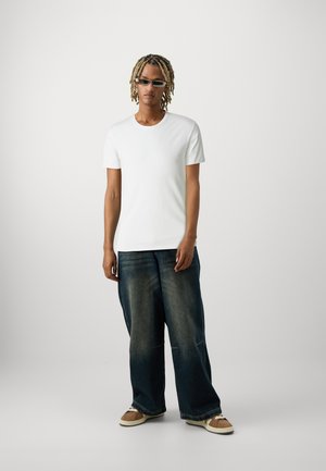 T-shirt en coton blanc, jean en denim bleu foncé à coupe ample avec décoloration, et baskets beiges avec semelles blanches. Accessoirisé avec des lunettes de soleil rectangulaires.