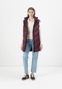 Veste rembourrée bordeaux avec un col haut, fermeture éclair à l'avant et poches latérales. Portée sur un haut blanc à côtes, associée à un jean bleu clair et des baskets noires.
