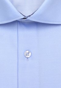 Gros plan d’un col de chemise habillée bleu clair avec un seul bouton blanc fixé sur la patte de boutonnage avant.