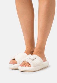 White fuzzy slippers with an adjustable strap and silver hardware, featuring a soft texture and a flat, light-colored sole.