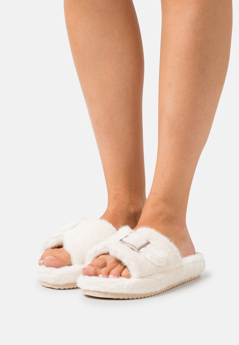 White fuzzy slippers with an adjustable strap and silver hardware, featuring a soft texture and a flat, light-colored sole.