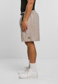 Beige Shorts aus leichtem Stoff, mit elastischem Bund, Seitentaschen und einem gelben Smiley-Logo am unteren Saum.
