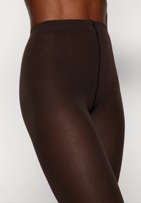 Leggings bruns transparents avec une texture lisse et une large ceinture élastique, présentant une couture centrale à l'avant pour un détail de design supplémentaire.