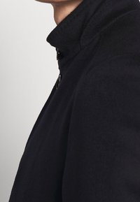 Cappotto di lana navy con colletto alto, texture liscia e dettagli dei bottoni ben visibili. Un design strutturato che enfatizza il collo e le spalle.