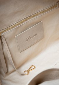 Beige handtasinterieur met een gouden rits, zachte stof, logo label en een gouden sluiting. Heeft een eenvoudig en strak ontwerp.