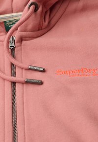 Sudadera con capucha rosa con cremallera y cordones con extremos metálicos grabados con "Superdry" y logo bordado naranja "SuperDry Athletics" en el pecho.