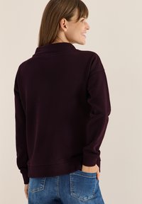 Felpa viola scuro con colletto alto, dotata di polsini e orlo a costine, indossata sopra jeans blu. Visibile la texture morbida e il taglio rilassato.
