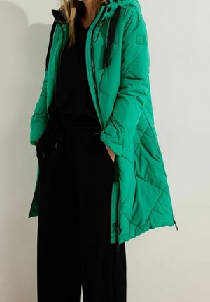 Femme portant un long manteau matelassé vert avec des cordons noirs et un pantalon noir décontracté, debout les mains dans les poches sur un fond uni.