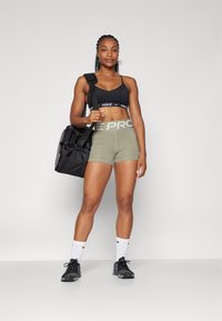 Černá sportovní podprsenka Nike, olivové kraťasy s vysokým pasem se textem "NIKE PRO", černá sportovní taška, bílé ponožky Nike a černé sportovní boty.