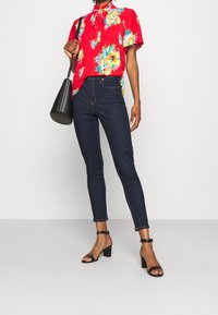 Röd blommönstrad blus med korta ärmar, kombinerad med mörkblå skinny jeans och svarta sandaler med blockklack och öppet tåparti. Svart handväska.