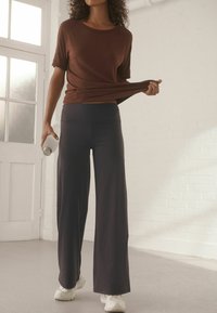 Brūns īsām piedurknēm blūze, brīvā griezumā, apvienota ar tumšiem platiem biksēm. Rokās turot baltu ūdens pudeli, ar baltām kedām. Minimalistisks fons.