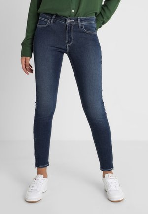 Jean slim fit bleu foncé porté avec des baskets blanches et une chemise verte à manches longues, debout devant un fond clair uni.