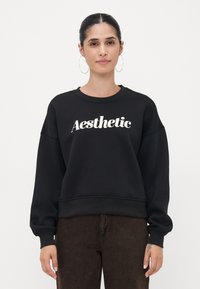 Felpa nera con collo rotondo e maniche lunghe, con la parola "Aesthetic" in bianco scritto in corsivo sulla parte anteriore.