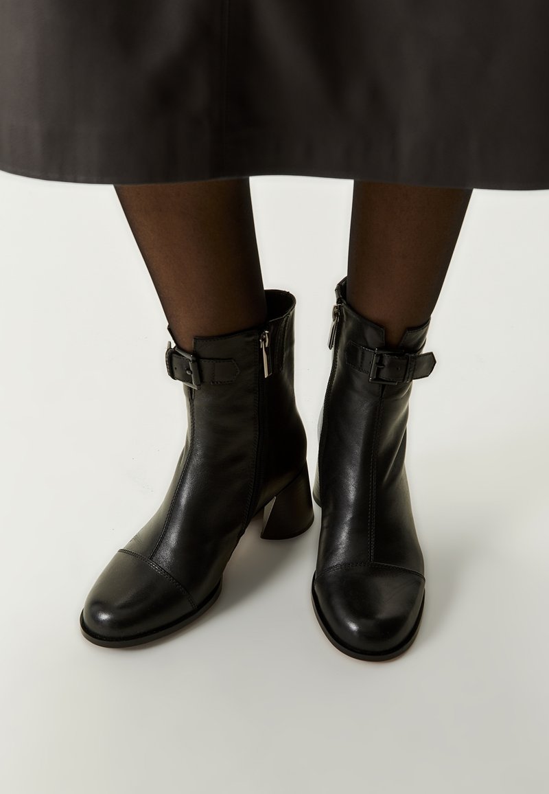 Botas de cuero negro hasta el tobillo con puntera cuadrada, cierre de cremallera y detalles decorativos de hebilla a los lados. Tacón cuadrado y textura suave.