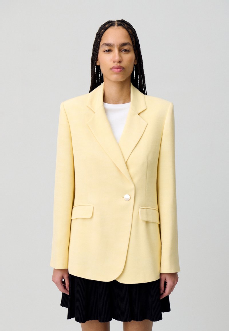 Blazer giallo con rever a punta, chiusura a un bottone e due tasche frontali. Tessuto testurizzato abbinato a un top bianco e una gonna a pieghe nera.