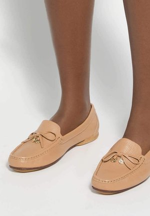 Feet wearing beige leather loafers with bow and gold charm details, on a plain white background.
