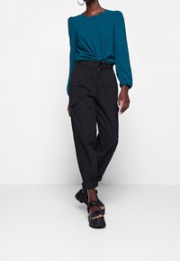 Maglia lunga teal con spalle a palloncino, annodata in vita, abbinata a pantaloni cargo neri larghi con tasche laterali e fondo con risvolto.