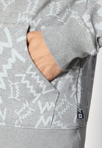 Hånd i lommen på en lysegrå sweatshirt med hvide abstrakte zigzag-mønstre og ribbede manchetter.