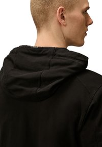 Zwarte hoodie met zichtbare naden; zachte stof; ontspannen pasvorm; voorzien van een trekkoordkap en een klein logo-etiket bij de nek.