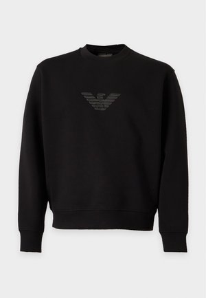 Homme portant une casquette noire, un sweat-shirt noir à manches longues avec logo, un jean bleu clair large et des baskets noires, debout sur fond blanc.
