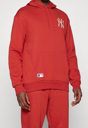 Man draagt een rode capuchontrui en bijpassende broek met het New York Yankees-logo op de borst en het MLB-logo op de zak.