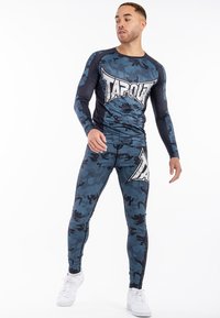 Hosszú ujjú kék terepmintás sportpóló, rajta a "TAPOUT" logóval, hozzá illő, testhez simuló leggingszel, mindkettő rugalmas anyagból készült. Fehér sportcipő.