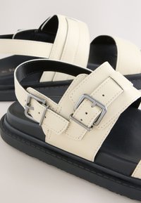 Sandales en cuir beige avec semelles intérieures noires rembourrées, dotées de boucles argentées réglables et de semelles épaisses en caoutchouc noir.