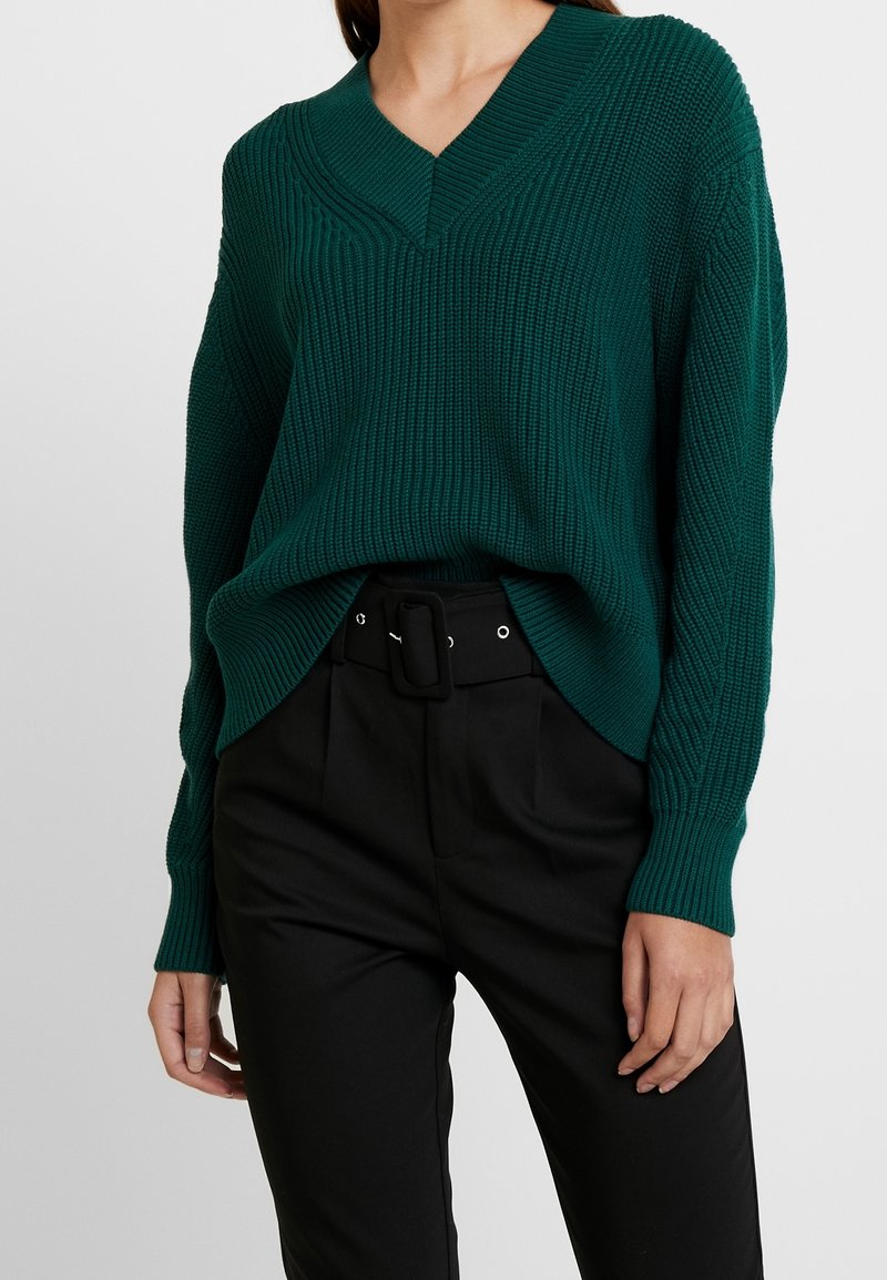 Femme portant un pull côtelé vert foncé à col en V, rentré dans un pantalon noir taille haute avec ceinture et boucle rectangulaire.
