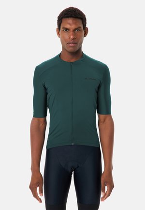 Homme athlétique portant un maillot de cyclisme vert foncé ajusté avec une fermeture éclair frontale et un cuissard de cyclisme noir rembourré, debout devant un fond uni.