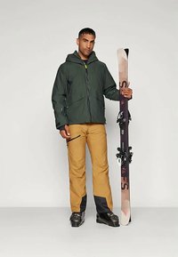 Mees, kellel on seljas roheline jakk, sinepikollased suusapüksid ja mustad suusasaapad, hoiab käes ühte vertikaalselt seismas olevat suuska koos sidemetega lihtsa taustaga.