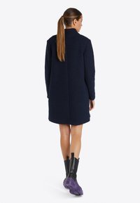 Navykék, oversized kabát texturált anyagból, álló gallérral, hosszú ujjakkal és egyenes szegéllyel, fekete és lila csizmával párosítva.
