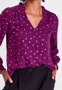 Blouse violette à col en V avec un décolleté à volants, manches longues avec poignets froncés, et un motif géométrique argenté. Fabriquée en tissu lisse.